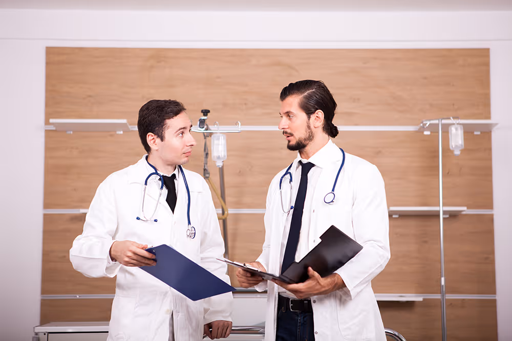 Two male doctors in white lab coats and ties, wearing stethoscopes, are standing and discussing documents on clipboards in a modern medical setting, symbolizing the need for practice leaders to collaborate on financial strategies.