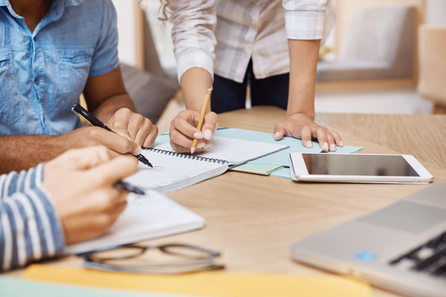 Close-up of people writing and sketching in notebooks on a wooden table with a tablet and laptop nearby.