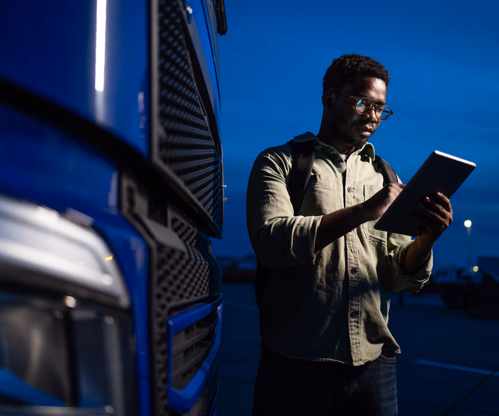 A logistics professional reviewing information on a tablet in a truck yard at dusk.