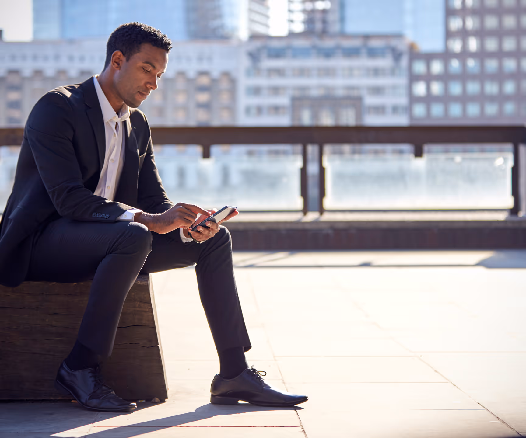 A man in a formal suit using technology, focused on his smartphone in a bright outdoor space.