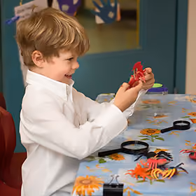 Young boy with light brown hair wearing a white shirt, smiling and holding a red toy crab while sitting at a table covered with a sunflower-patterned tablecloth and various plastic sea creatures and magnifying glasses.