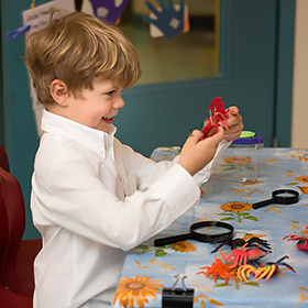 Young boy with light brown hair wearing a white shirt, smiling and holding a red toy crab while sitting at a table covered with a sunflower-patterned tablecloth and various plastic sea creatures and magnifying glasses.