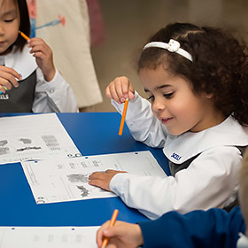 Young children in school uniforms sitting at a blue table, drawing and coloring on worksheets with orange pencils.