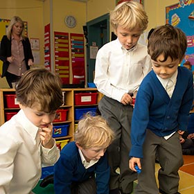 Four young boys in school uniforms engaged in an activity on the floor in a classroom, with a female teacher observing in the background.
