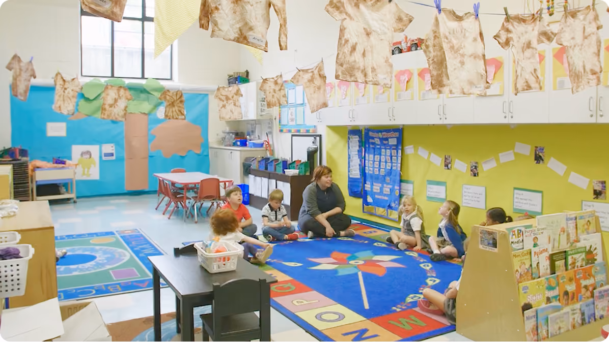 A preschool classroom with children and a teacher sitting on a colourful alphabet rug, surrounded by educational materials and clothes hanging above.
