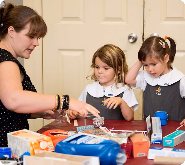 A female teacher with bracelets on her wrist instructs two young girls in school uniforms seated at a table covered with various craft supplies and materials in a classroom setting.