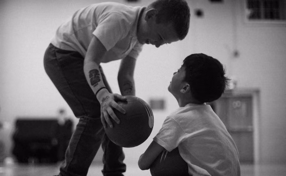 Black and white photo of an adult and a child in a gymnasium where the adult leans down holding a basketball, and the child sits on the floor looking up at them.
