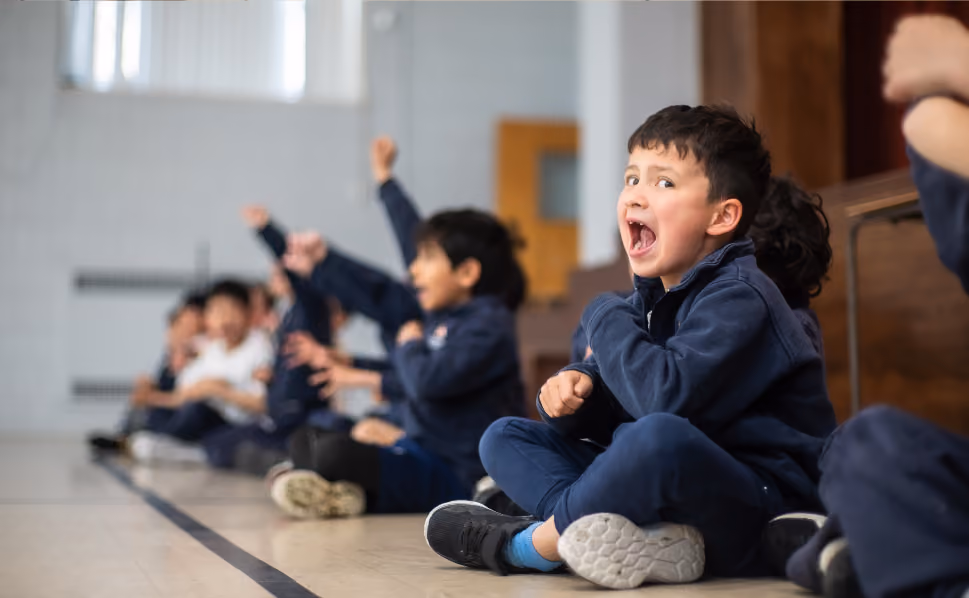 A group of children sitting on the floor indoors, with one boy in the foreground enthusiastically raising his fist and shouting.