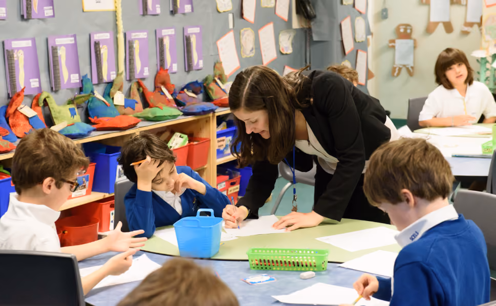 Teacher leaning over a table helping young students with their schoolwork in a colourful classroom filled with student projects on the walls and shelves.