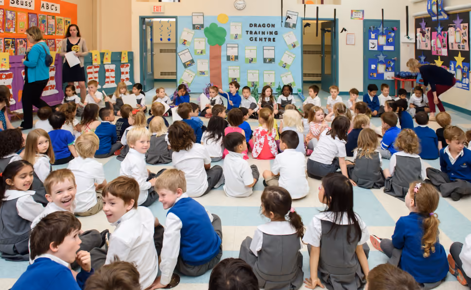 Large group of young school children in uniforms sitting on the floor facing a bulletin board labeled Dragon Training Centre in a classroom with educational displays and teachers present.