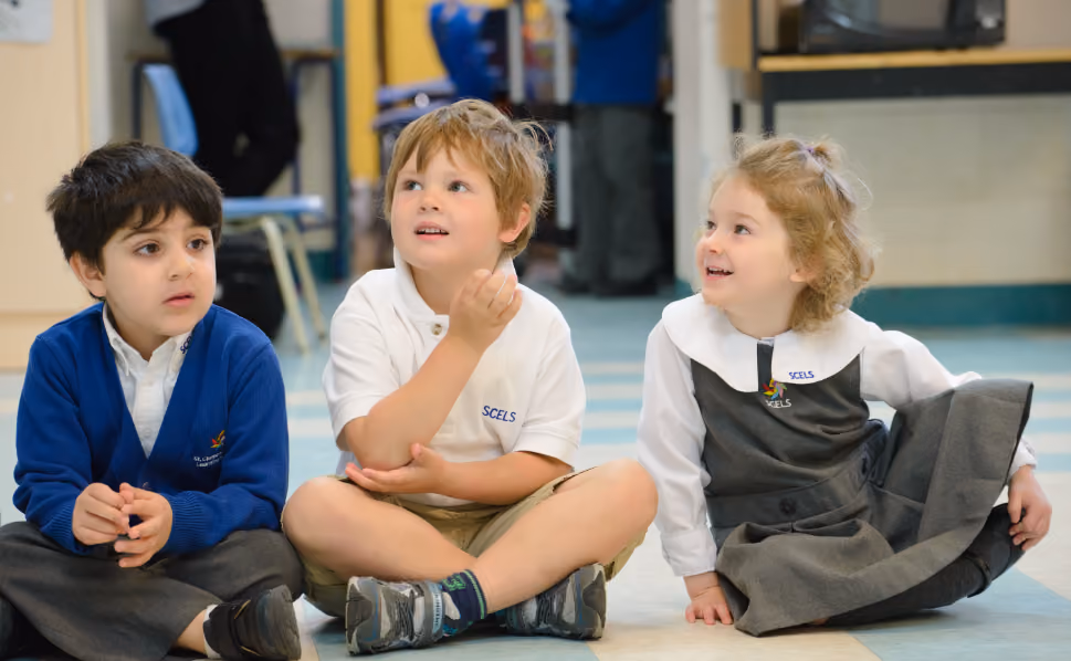 Three young children sitting on a classroom floor, two boys and one girl, wearing school uniforms with the SCELS logo, engaged and looking slightly upward.