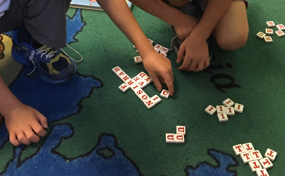 Two children sitting on a colorful map-patterned carpet playing with letter tiles that spell out words including 'NOSTRA' and 'LIBERTAD'.