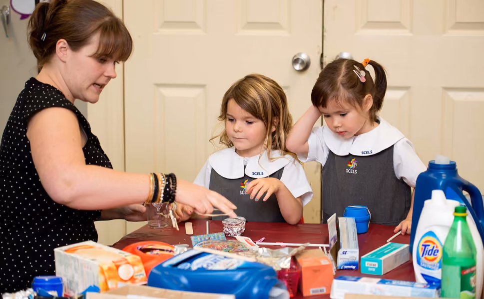 A woman with bracelets is instructing two young girls in school uniforms at a table filled with various household products including laundry detergent, soap, and boxes, as they look attentively at the items in front of them.