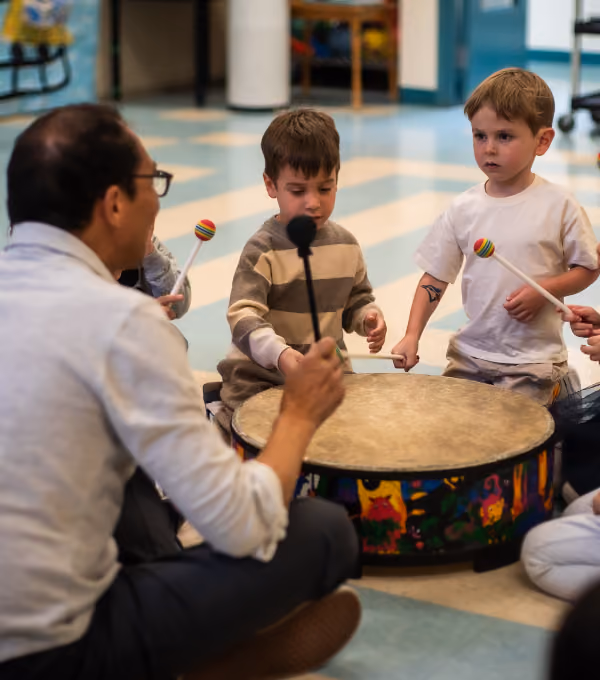 Adult and three children sitting around a large colourful drum, each holding mallets with colourful round heads, playing the drum indoors on a tiled floor.