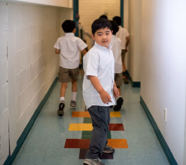 Young boys in school uniforms walking and playing hopscotch in a brightly lit school hallway with tiled floor.