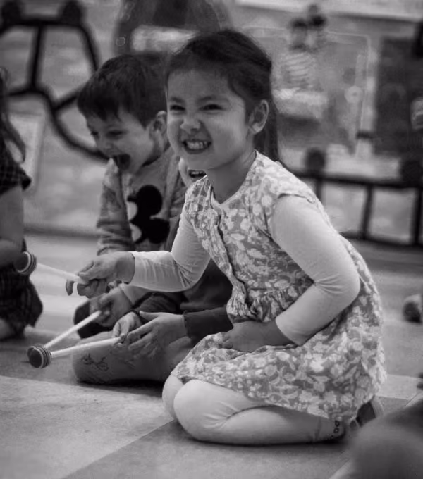 A smiling young girl kneels on the floor playing percussion mallets in a group activity with other children.