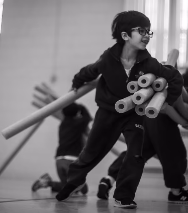 Black and white photo of a young boy wearing glasses and a dark outfit running indoors while carrying several foam pool noodles under one arm.