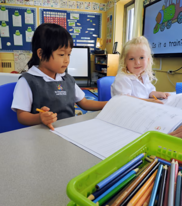Two young girls sitting at a classroom table working on worksheets; one girl has dark hair and wears a school uniform, the other has blonde hair and wears a white shirt. Coloured pencils are in a green basket on the table, and a screen in the background shows a cartoon train and the text 'Is it a train?'.