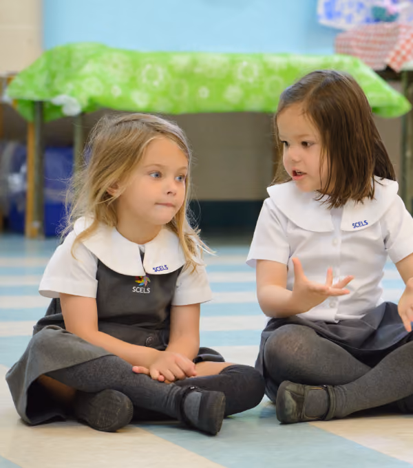 Two young girls sitting cross-legged on a tiled floor inside a room; both wear matching school uniforms with white collars and dark skirts, and appear to be engaged in conversation.