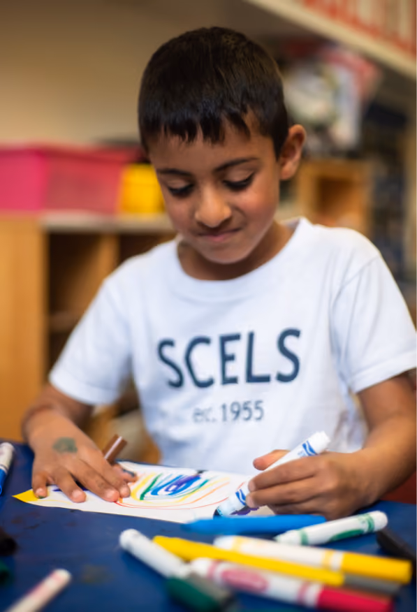 Young boy with dark hair wearing a white SCELS t-shirt focused on coloring a paper with blue, green, and yellow markers at a table with scattered markers.