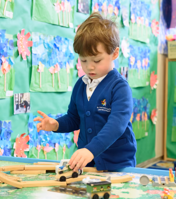 A young boy wearing a blue St. Clement's Early Learning School cardigan plays with toy vehicles and wooden tracks in a colourful classroom with children's artwork on the walls.