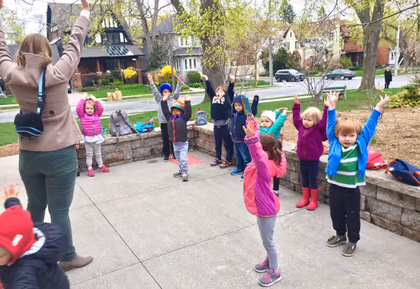 A group of children standing outdoors in a semi-circle on a paved area with their arms raised, following the lead of an adult who also has raised arms, with houses and trees in the background.