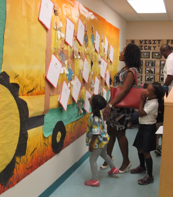 A woman and two young girls look at a classroom bulletin board decorated with yellow and orange background and children's drawings or writings pinned in organized rows.