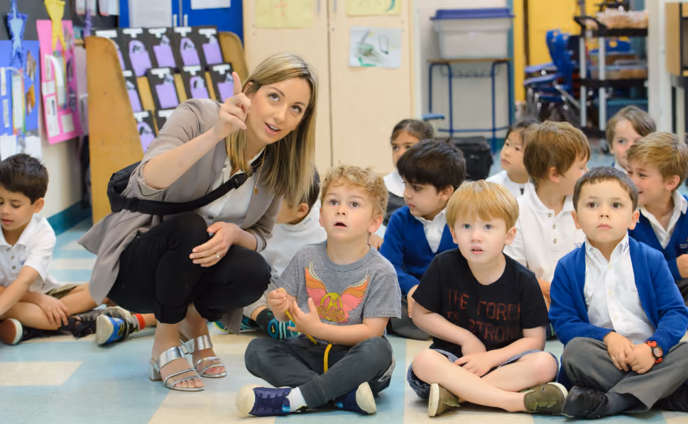 A female teacher crouching and pointing while speaking to a group of young children sitting on the classroom floor, with colourful children's artwork displayed on the walls behind them.