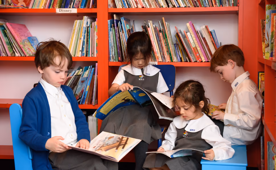 Four young children in school uniforms reading books while seated in front of orange bookshelves filled with colorful books.