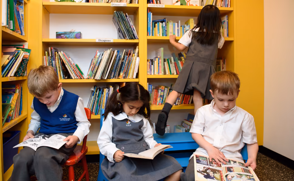 Three children sitting and reading books in front of yellow library shelves filled with books, while a fourth child climbs a blue step stool to reach higher shelves.