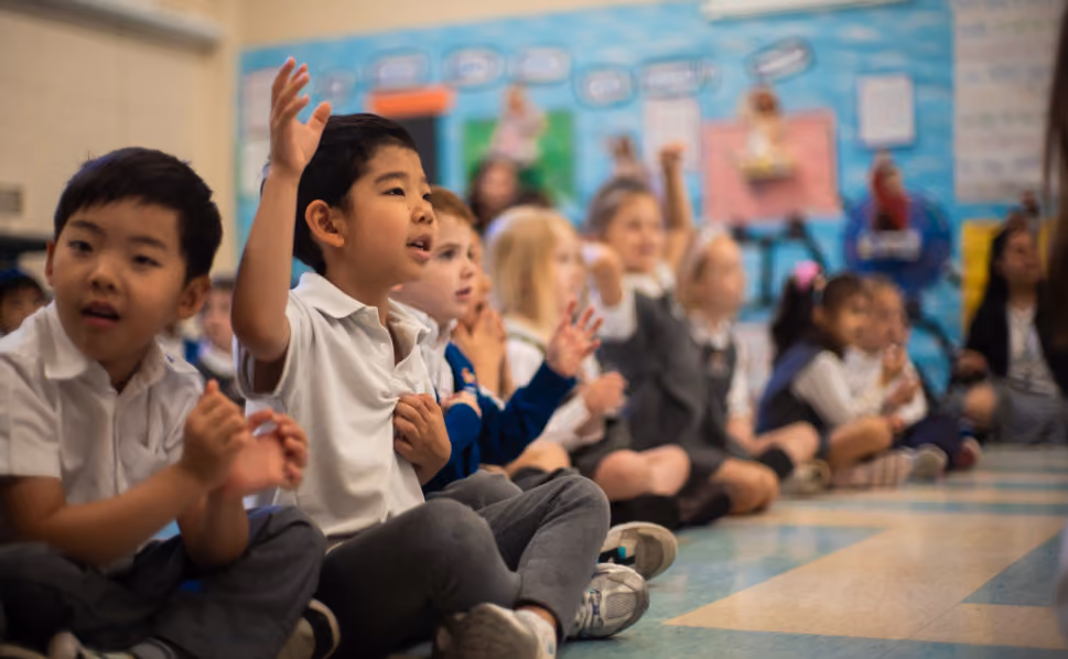 A group of diverse young schoolchildren sitting cross-legged on a classroom floor, some with raised hands, actively participating in a lesson.