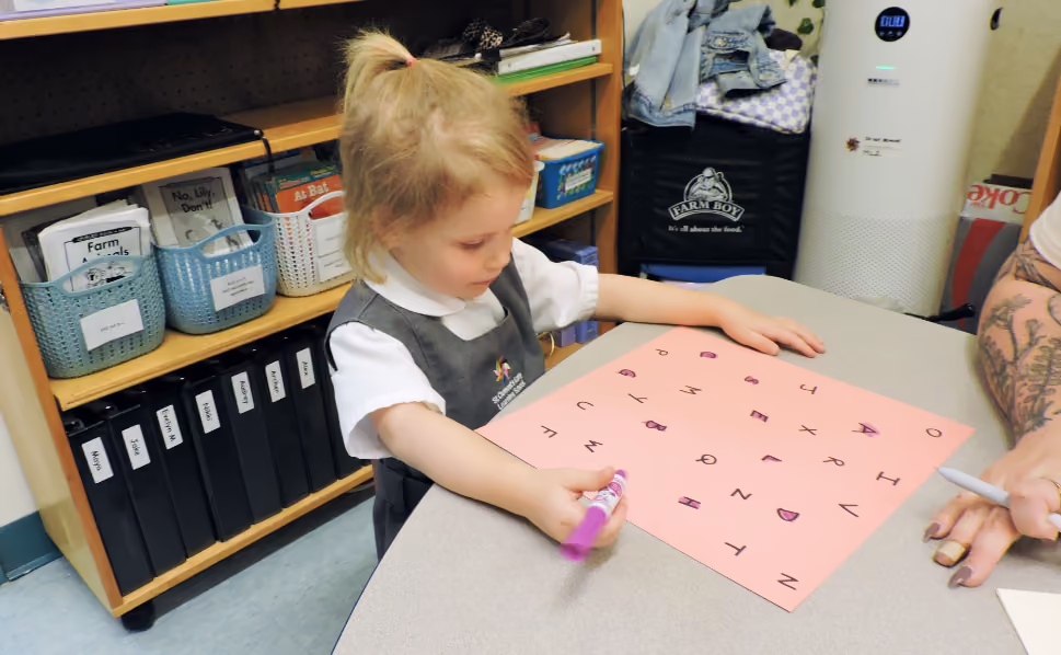 A young girl in a gray school uniform with a white shirt sits at a table and writes on a pink sheet of paper covered with handwritten uppercase letters using a purple marker, with a tattooed adult's hand nearby.