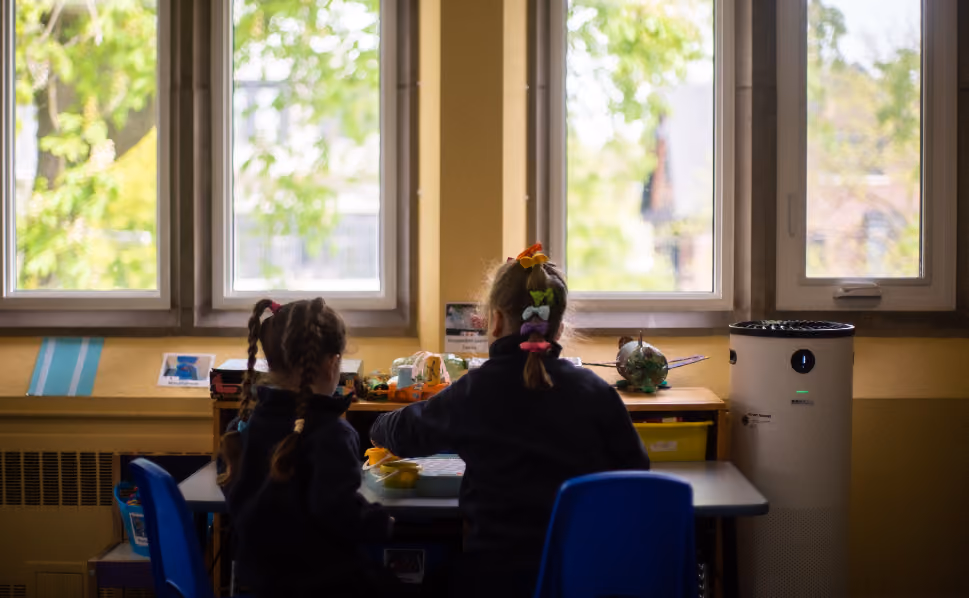 Two young children with braided hair sitting at a table facing a window with green trees outside, in a softly lit classroom setting with toys and an air purifier nearby.