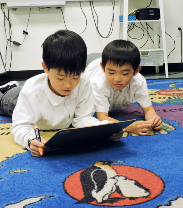 Two young boys lying on a colorful classroom carpet with penguin designs, one boy holding a black clipboard and an orange marker while both focus on the board.