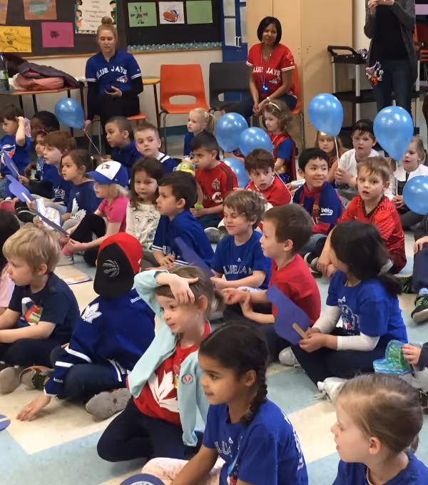 Group of young children sitting on a classroom floor, many wearing Blue Jays shirts and holding blue balloons, with two adults in Blue Jays jerseys seated in the background.
