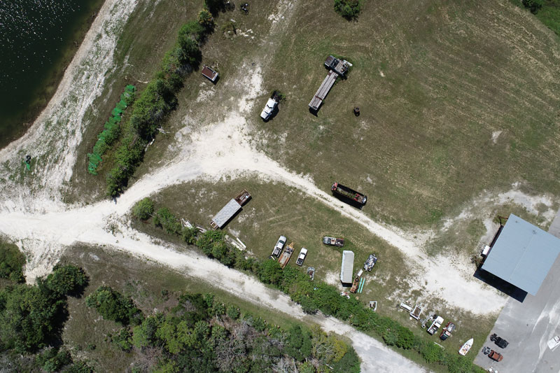 Aerial view of a rural waterfront property with gravel access roads, utility structures, open land, and shoreline access.