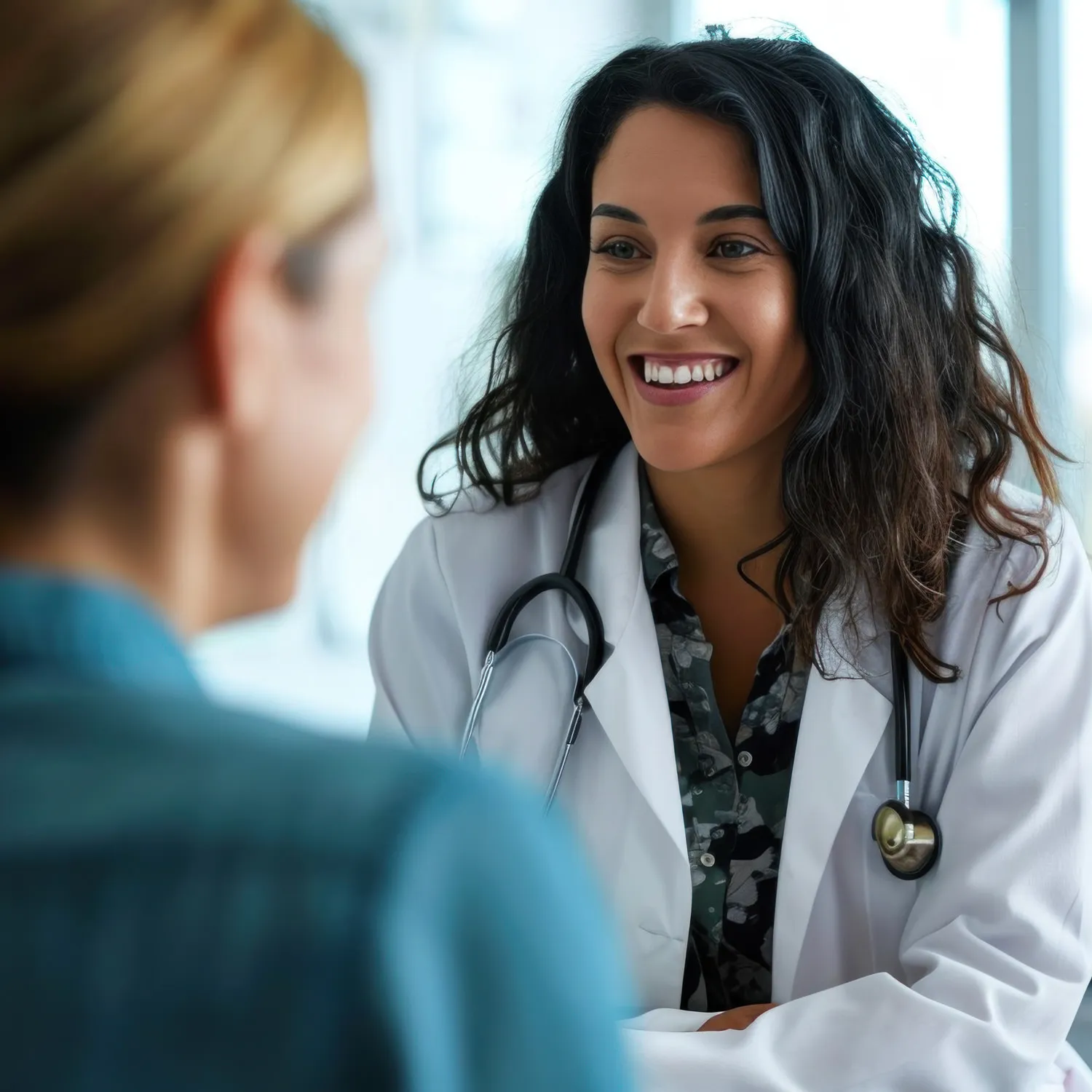 Smiling female doctor wearing a white coat and stethoscope talking to a patient in a medical office.