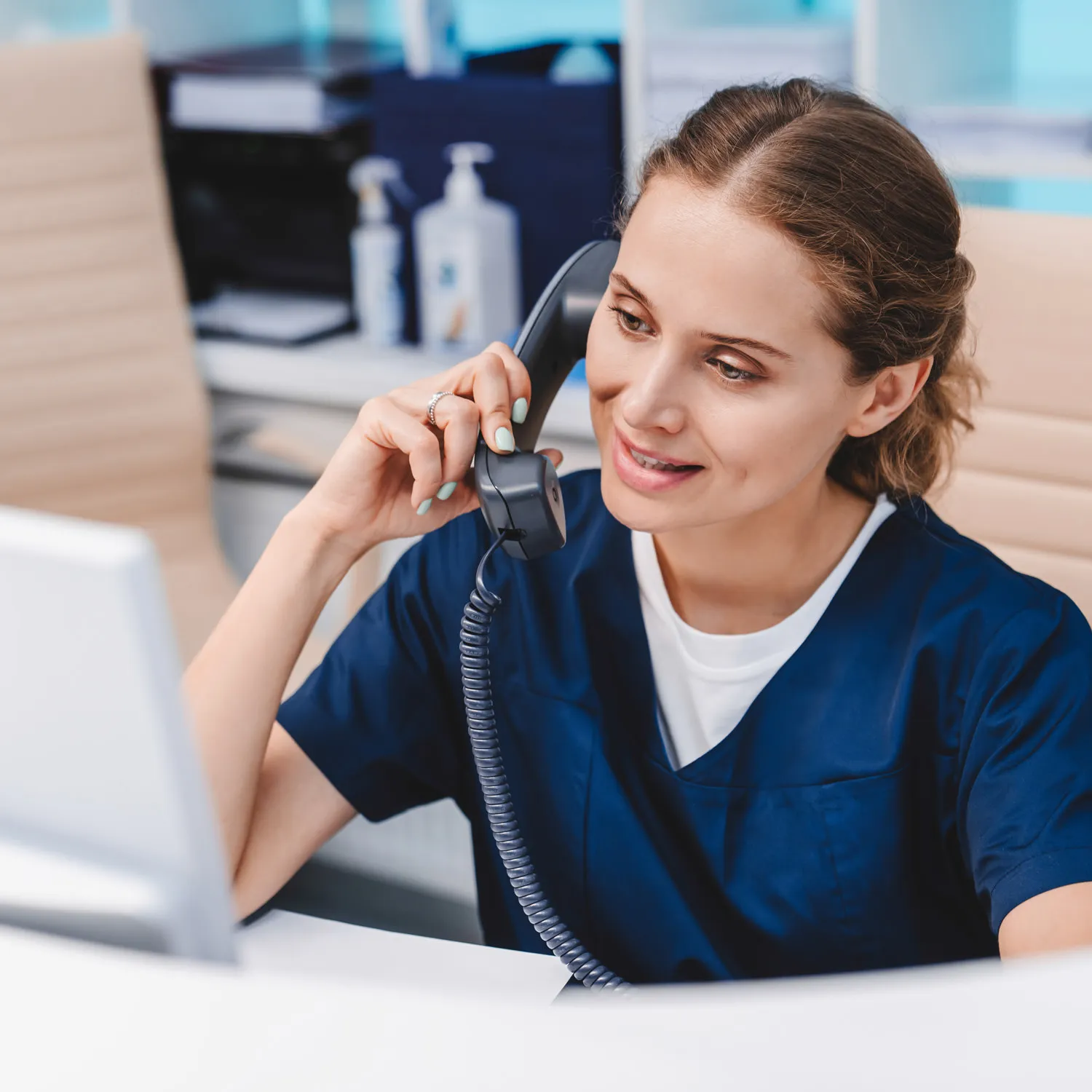 Female healthcare professional in navy scrubs talking on a corded office phone at a desk.