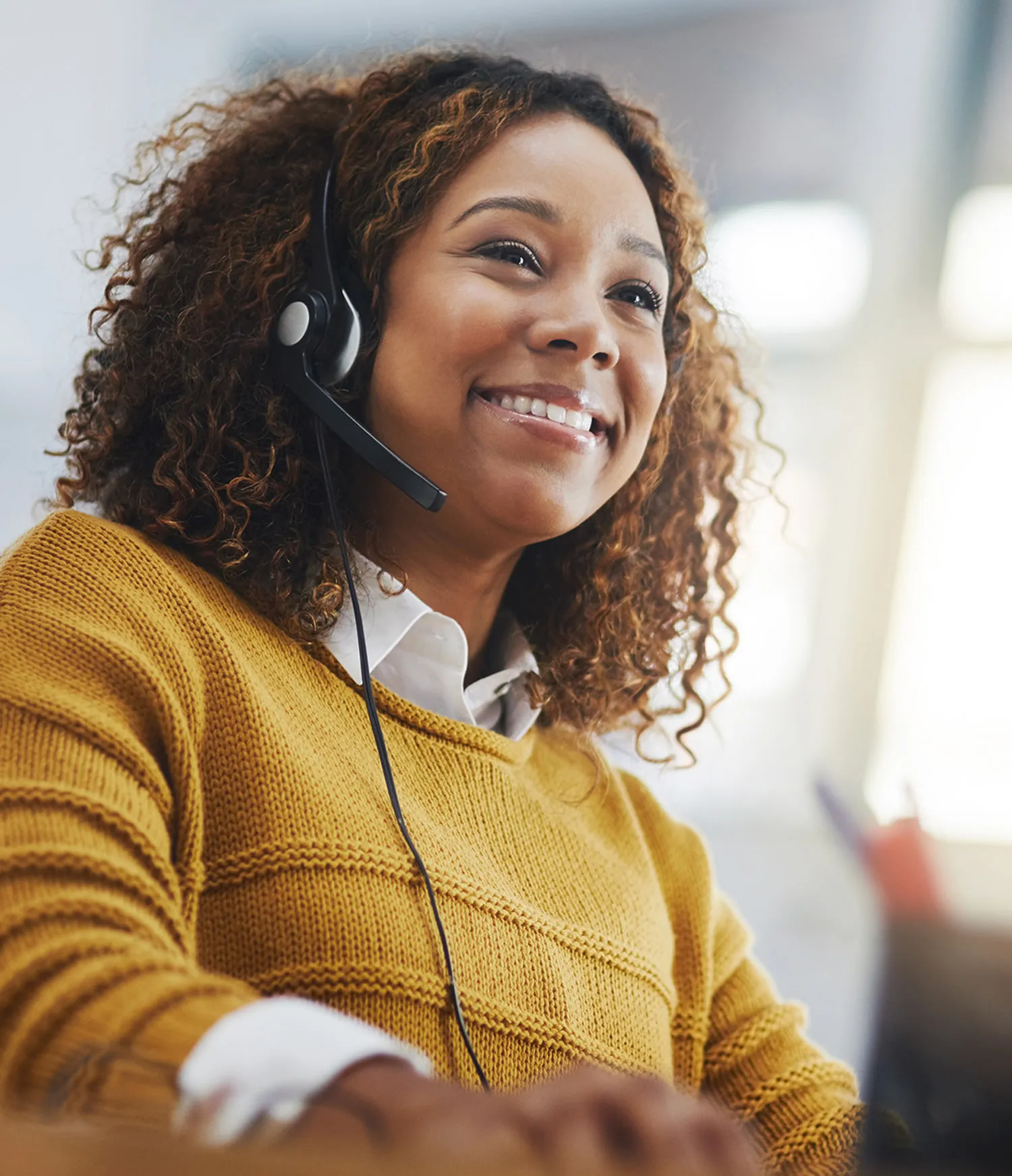 Smiling woman with curly hair wearing a headset and a mustard yellow sweater.