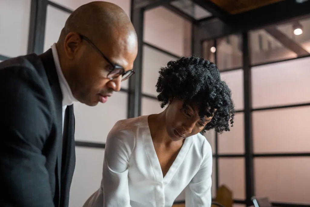 Two business professionals, a man wearing glasses and a woman with curly hair, attentively reviewing documents in an office.