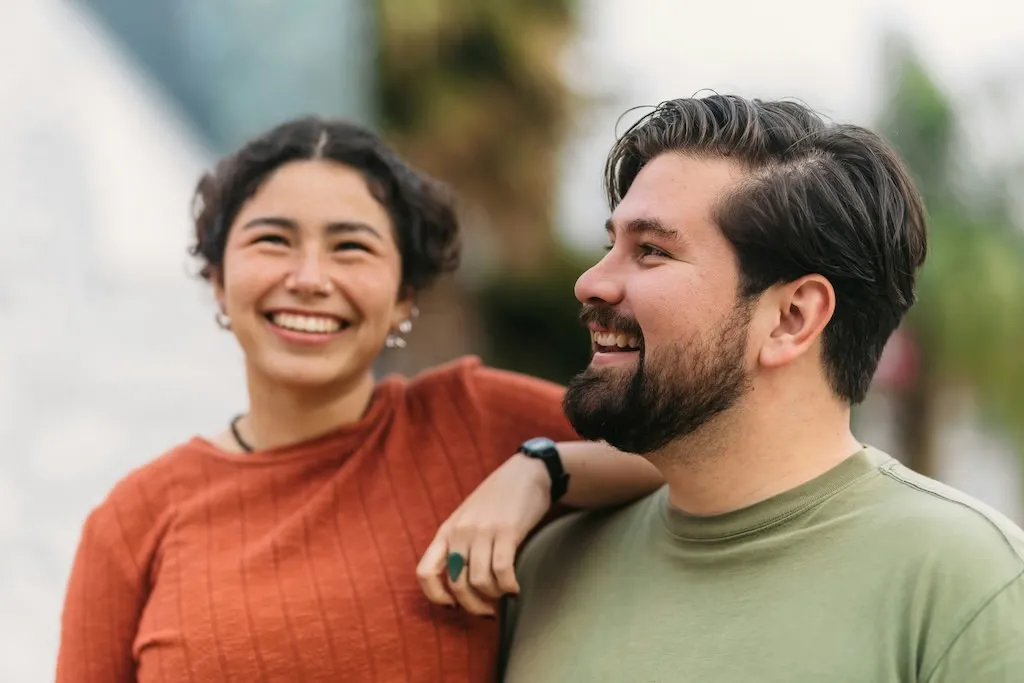 Young woman in an orange sweater smiling with her arm around a bearded man in a green shirt.