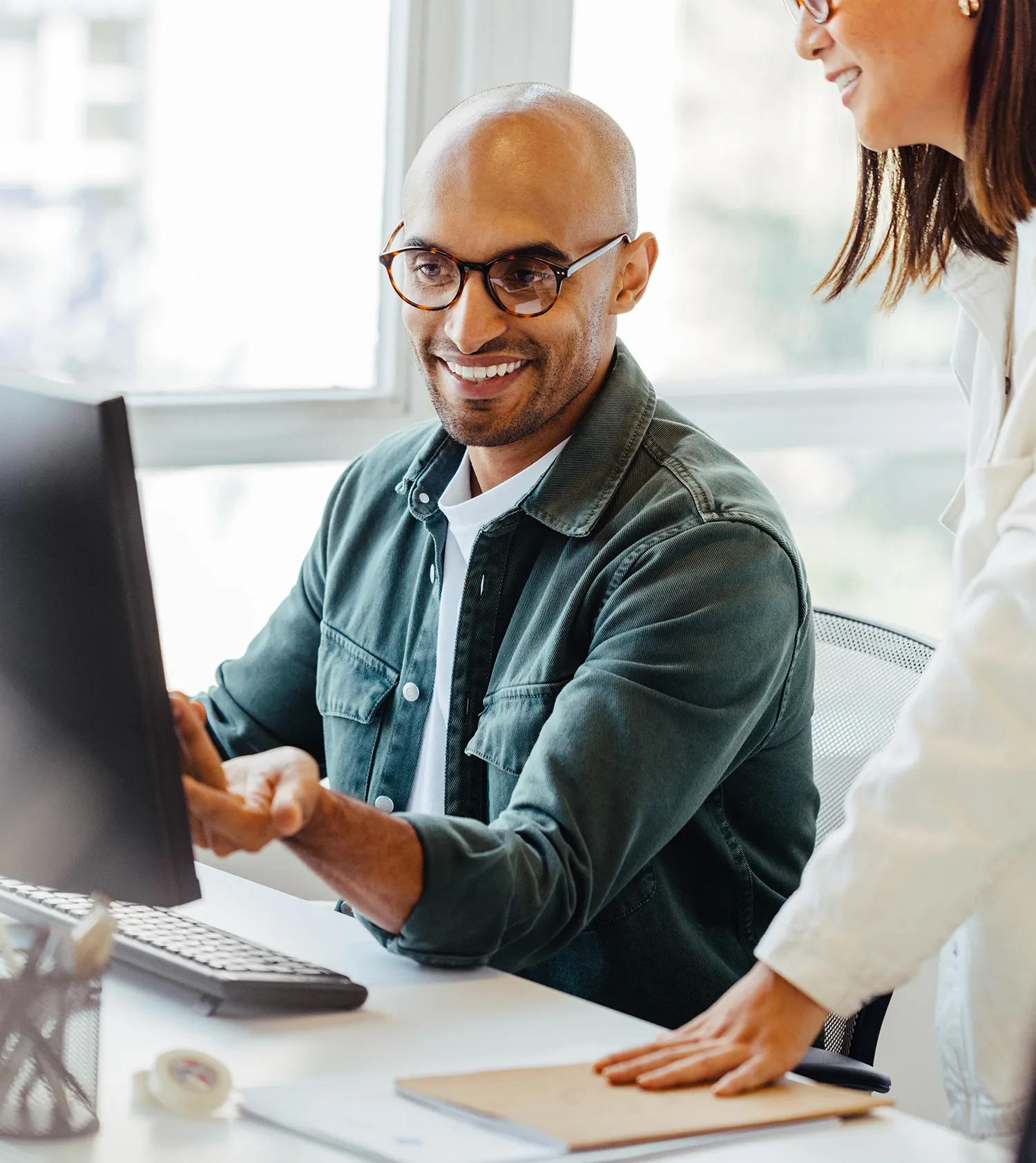 Smiling man wearing glasses pointing at a computer screen while a woman stands beside him in a bright office.