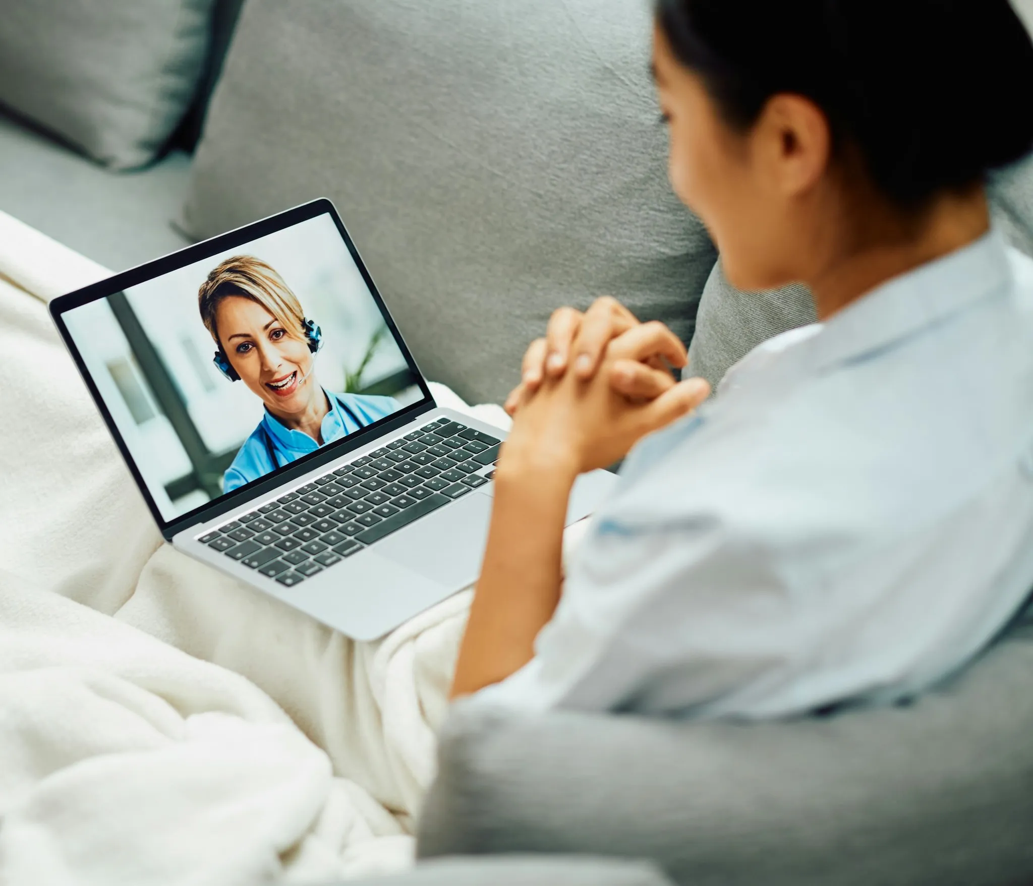 Woman sitting on a couch video calling a healthcare professional wearing a headset on a laptop.