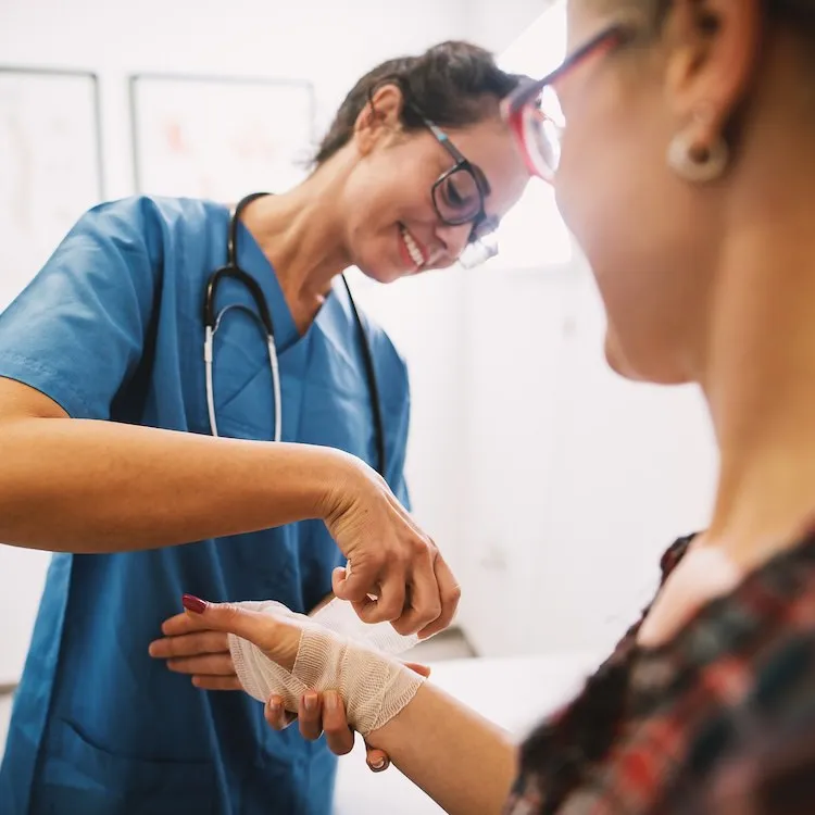 Healthcare professional in blue scrubs and glasses smiling while wrapping a bandage around a patient's hand.
