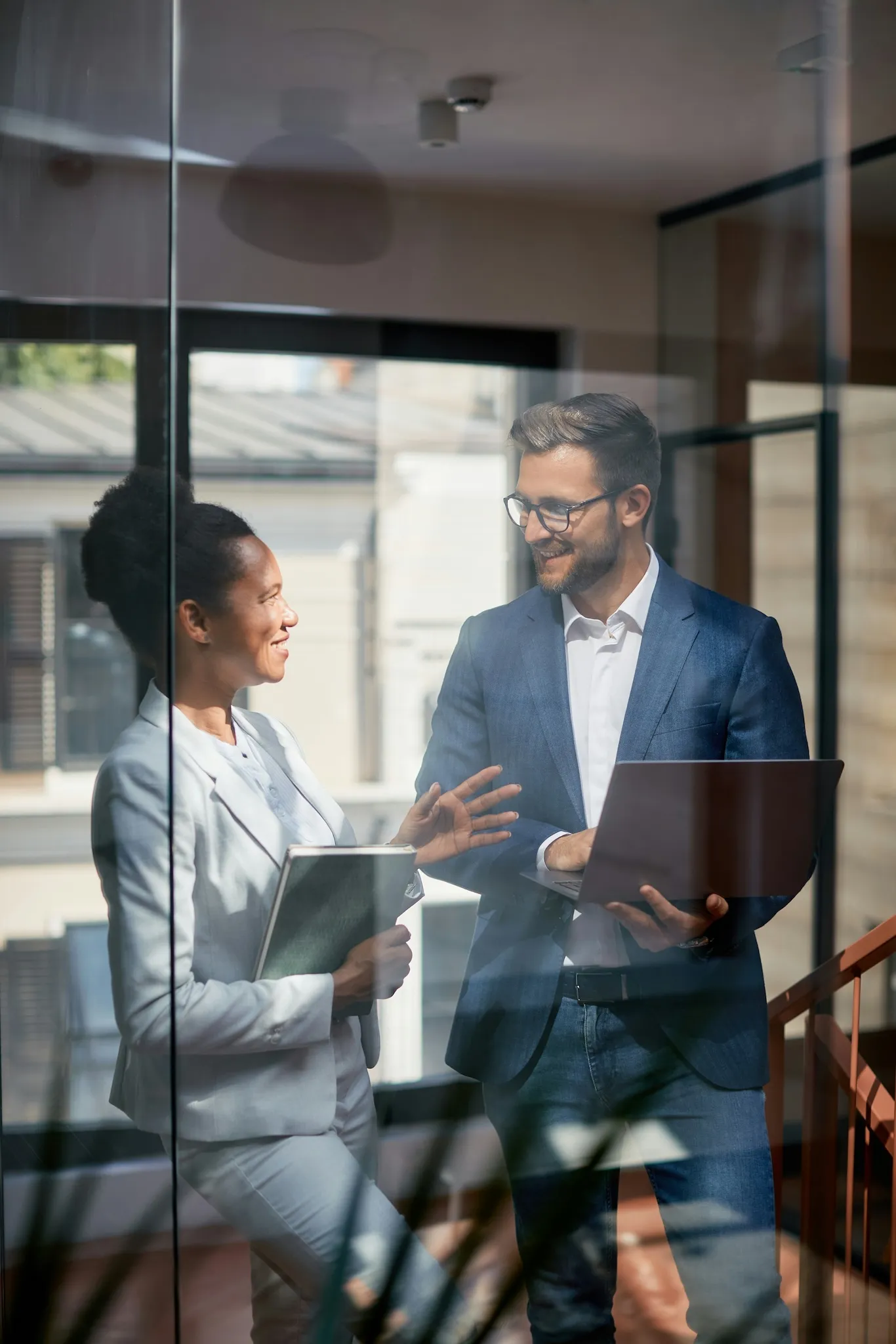 Two business professionals having a conversation indoors, one holding a laptop and the other holding a notebook.