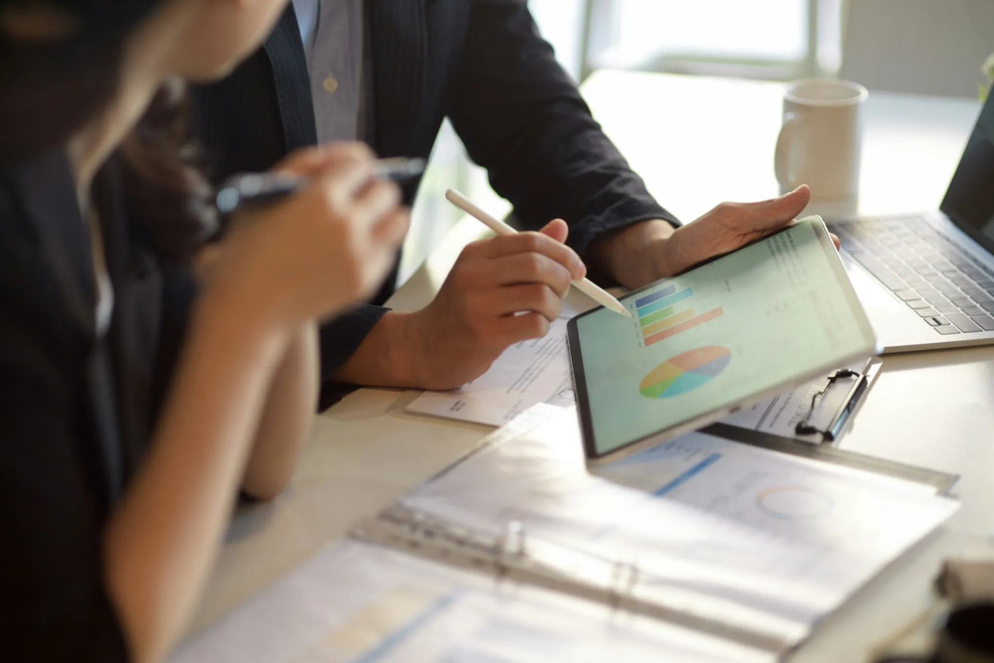 Two people reviewing colorful bar and pie charts on a tablet over a table with documents and a laptop.