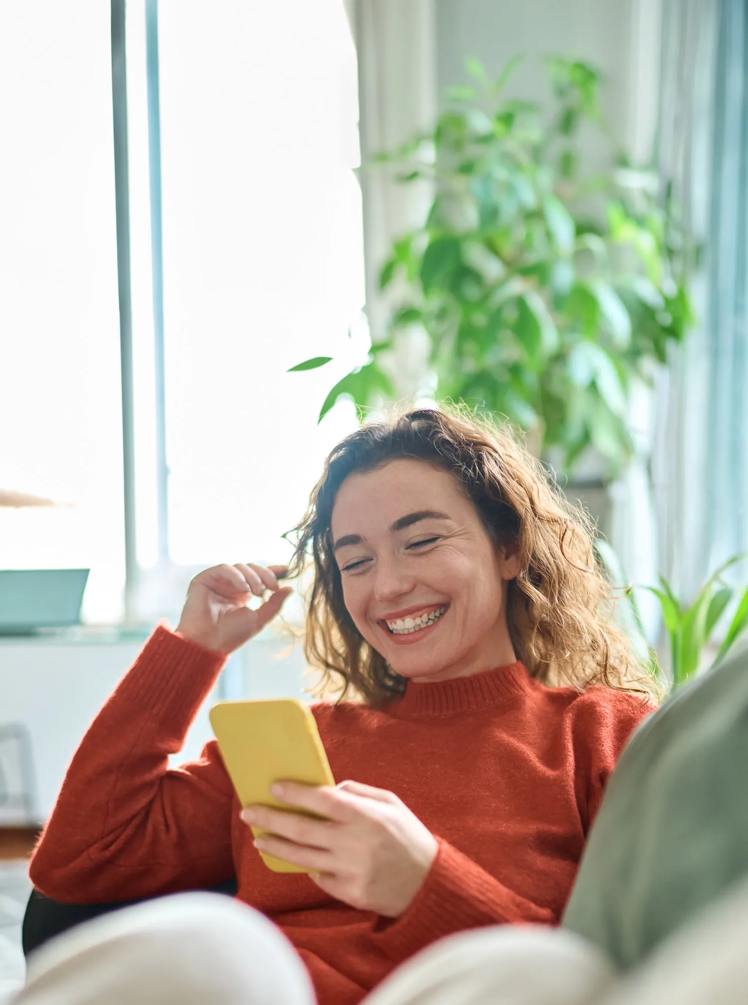 Smiling woman in a red sweater looking at a yellow smartphone in a bright room with plants.