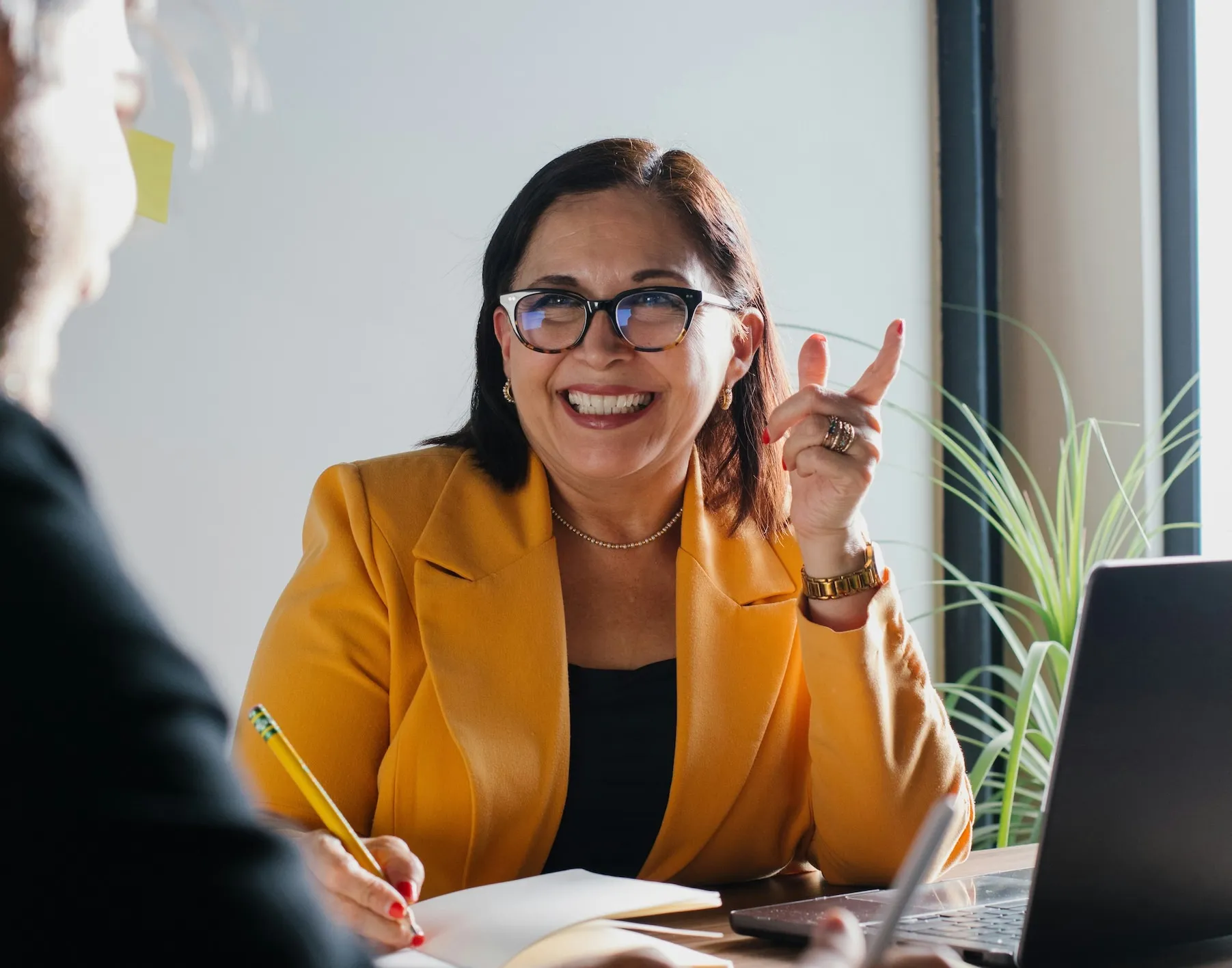 Smiling woman in a yellow blazer and glasses holding a pencil and gesturing while talking during a meeting.