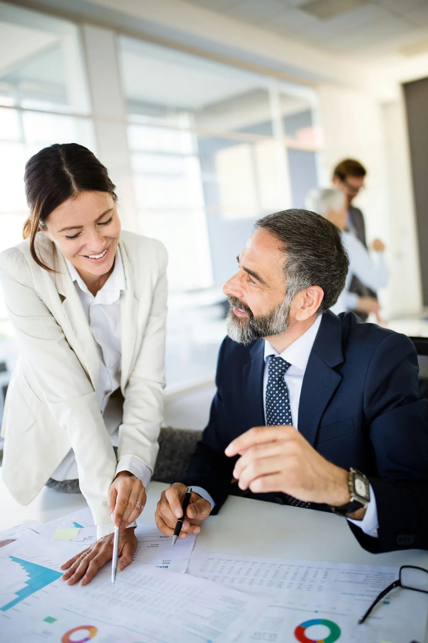 A man and woman in business attire discussing charts and documents at a desk in a bright office.