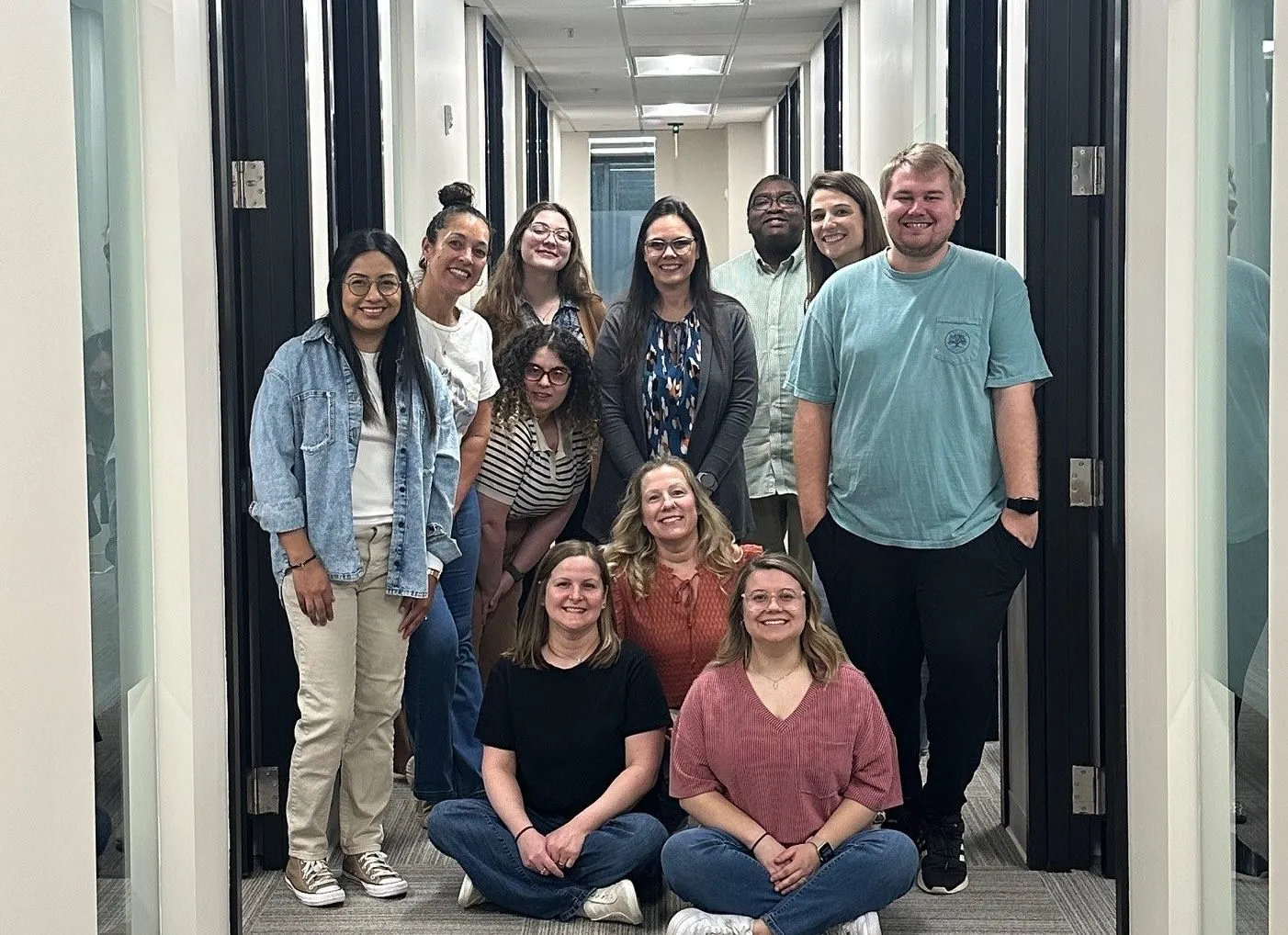 Group of eleven diverse people smiling and posing together in a hallway with office doors on both sides.